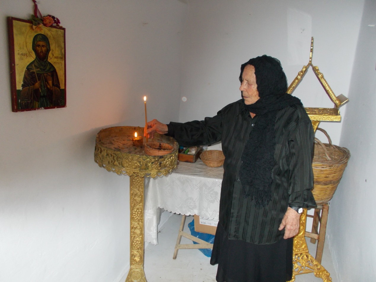 Η Ανδρονίκη Κουρμούλη ανάβει κερί στο εκκλησάκι του Αγίου Νικολάου του Κουρταλιώτη των Αγκουσελιανών στη μνήμη των θυμάτων των Σκουρβούλων και όλων των νεκρών της Κατοχής
